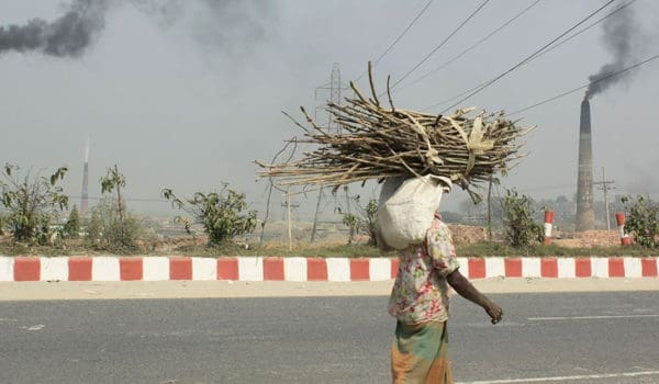 Fra Dhaka, Bangladesh. Mann bærer bør på hodet. Fabrikkpiper med svart røyk i bakgrunnen.