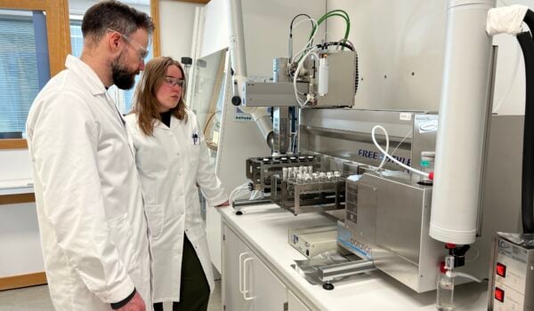 To forskere i hvite frakker står i et laboratorium foran en maskin som analyserer prøver for ulike kjemikalier.